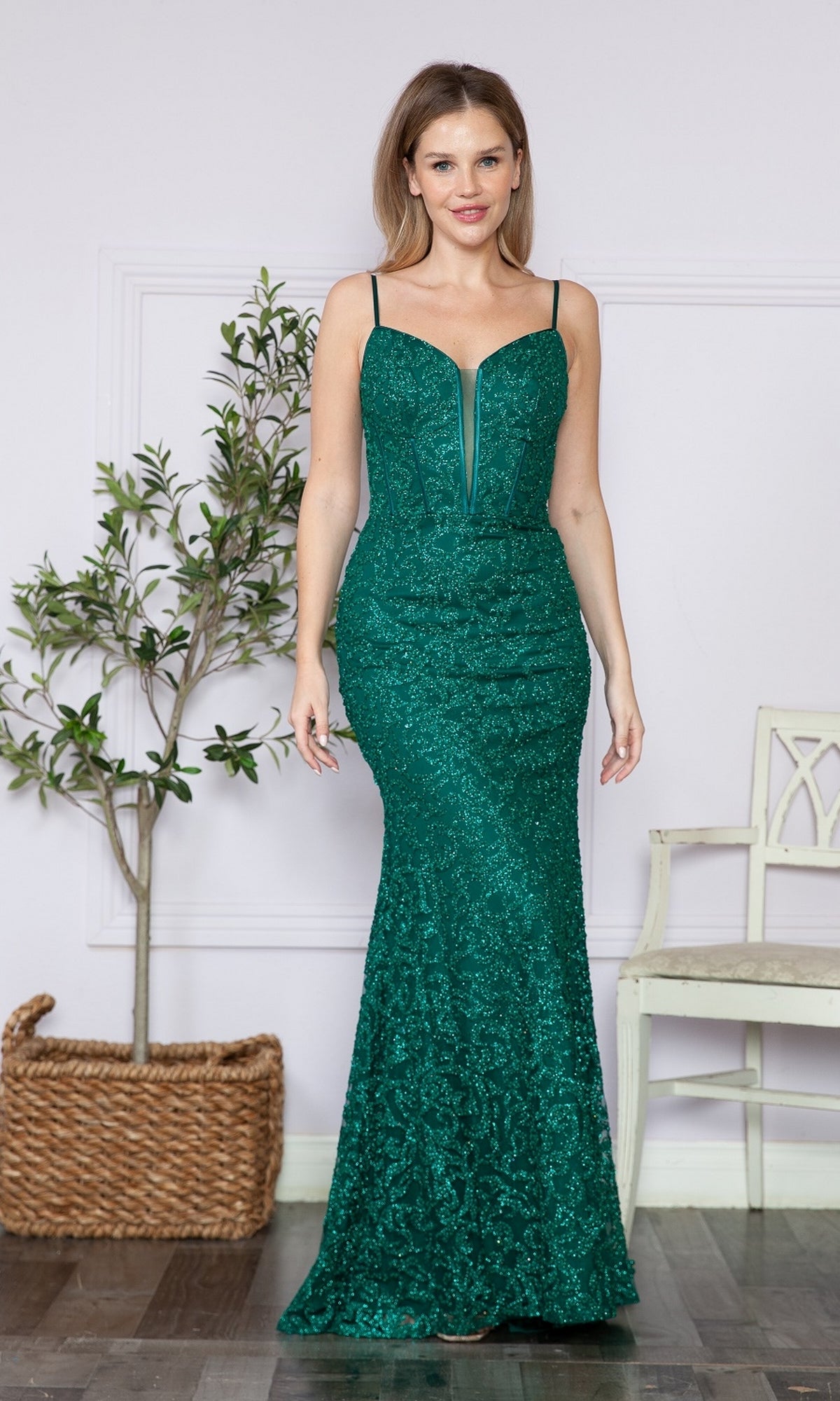 A woman stands indoors wearing the Glitter-Lace Long Corset Prom Dress 9354 in emerald, a sleeveless, floor-length gown with thin straps. A potted plant and a white chair are visible in the background.